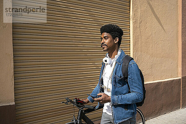Afro-Mann mit Fahrrad hält Smartphone an der Wand während eines sonnigen Tages