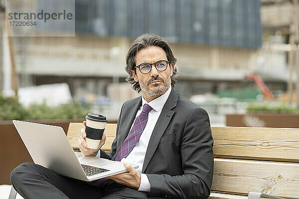 Nachdenklicher Geschäftsmann mit Kaffee in der Hand und Laptop auf einer Bank