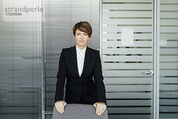 Geschäftsfrau mit ausdruckslosem Blick hinter einem Stuhl im Büro stehend