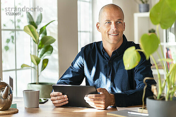 Lächelnder Geschäftsmann  der ein digitales Tablet hält  während er in seinem Büro wegschaut
