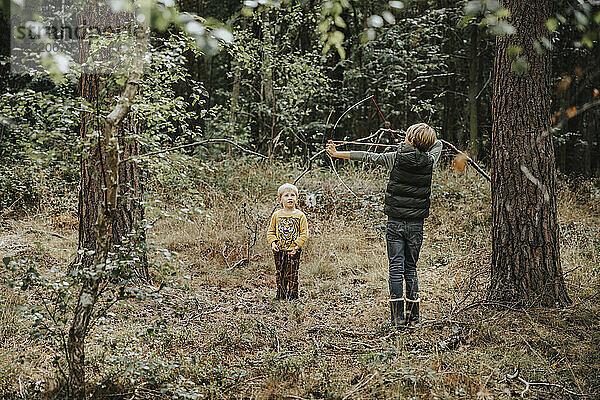 Junge spielt mit Pfeil und Bogen  während sein Bruder neben ihm im Wald steht