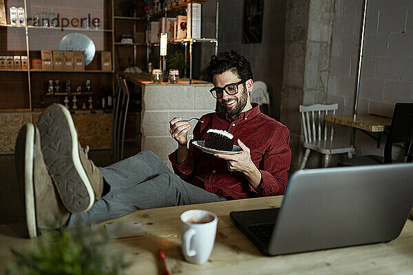 Lächelnder Geschäftsmann mit Laptop und Kaffee  der in einem Café ein Stück Kuchen isst