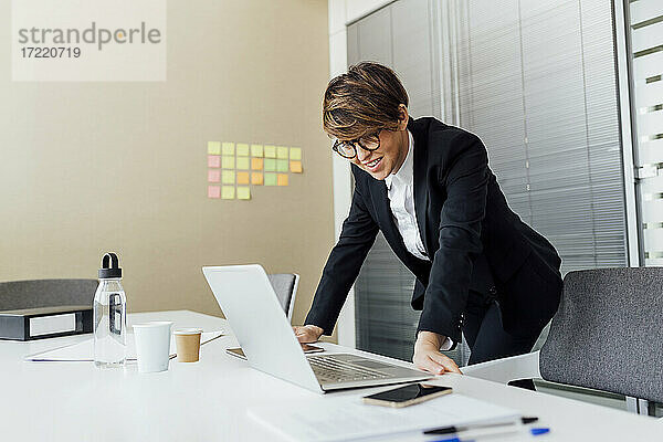 Lächelnde Geschäftsfrau  die auf einen Laptop schaut  während sie sich an einen Schreibtisch im Büro lehnt