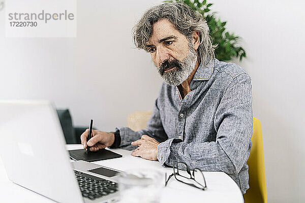 Älterer männlicher Geschäftsmann  der auf seinen Laptop schaut  während er im Büro zu Hause ein Grafiktablett benutzt