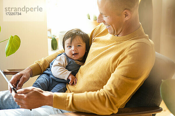 Vater hält digitales Tablet und lächelndes Baby  während er zu Hause auf einem Sessel sitzt