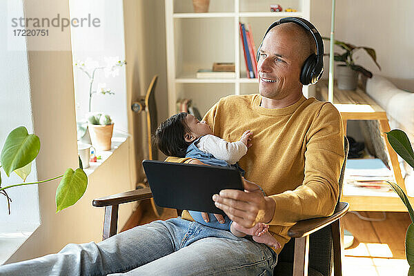 Lächelnder Vater hört Musik  während er ein schlafendes Baby und ein digitales Tablet im Wohnzimmer hält