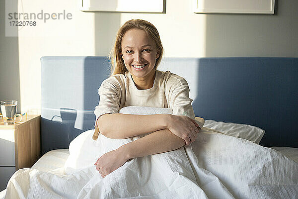 Junge Frau lächelt  während sie die Knie auf dem Bett zu Hause umarmt