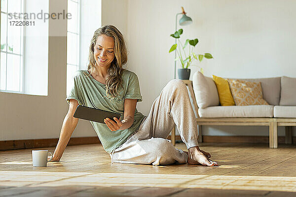 Lächelnde Frau schaut auf ein digitales Tablet  während sie auf dem Boden sitzt
