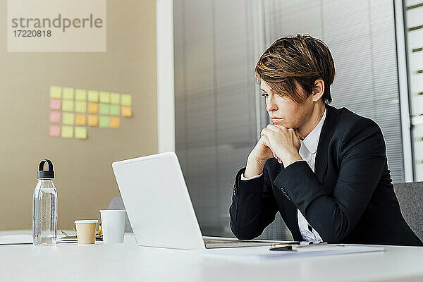 Nachdenkliche Unternehmerin mit Hand am Kinn  die auf ihren Laptop schaut  während sie im Büro sitzt