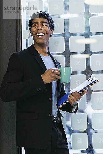 Lächelnder Geschäftsmann mit Kaffeetasse und Aktenordner  der am Fenster steht
