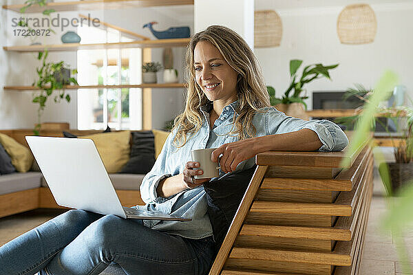 Lächelnde Frau mit Kaffeetasse  die ein Video auf einem Laptop im Wohnzimmer auf der Couch ansieht