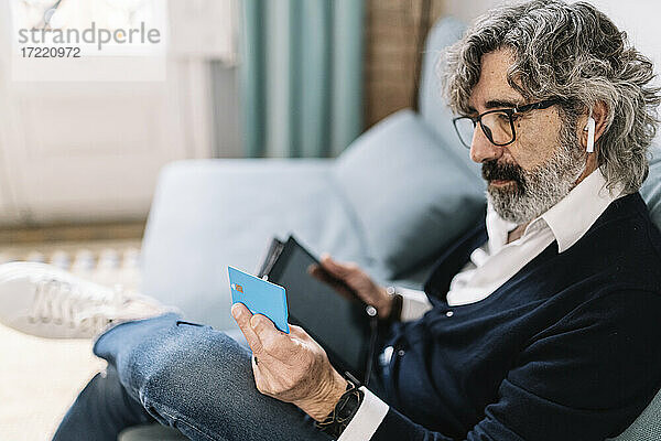Gut aussehender Mann  der eine Kreditkarte betrachtet und ein digitales Tablet hält  während er zu Hause sitzt