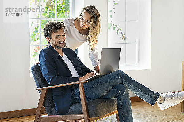 Lächelnde Freundin sieht ihren Freund an  der einen Laptop benutzt  während er zu Hause auf einem Stuhl sitzt