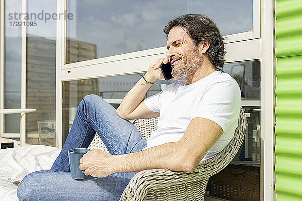 Mann hält Tasse und spricht mit seinem Smartphone  während er auf einem Sofa auf dem Balkon sitzt