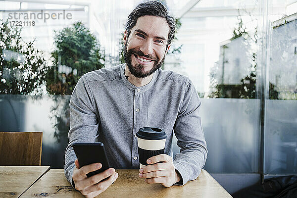 Glücklicher männlicher Geschäftsmann mit Smartphone und wiederverwendbarem Kaffeebecher im Büro