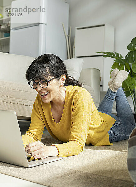 Fröhliche Frau mit Brille benutzt einen Laptop  während sie zu Hause auf dem Boden liegt