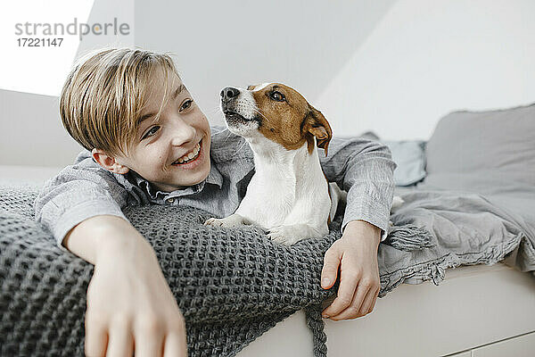 Lächelnder Junge mit Arm um den Hund  der zu Hause auf dem Bett liegt