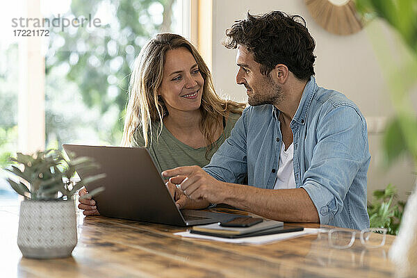 Lächelndes Paar  das sich gegenseitig ansieht  während es vor einem Laptop am Tisch sitzt