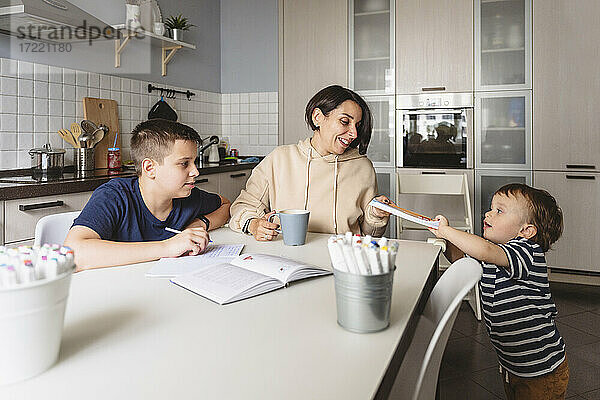 Junge gibt seiner Mutter ein Buch  während sein Bruder am Küchentisch Hausaufgaben macht