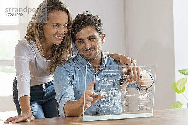Mid adult couple looking at floor plan over transparent screen at home