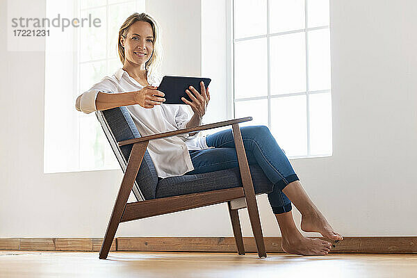 Lächelnde Frau mit digitalem Tablet auf einem Stuhl zu Hause