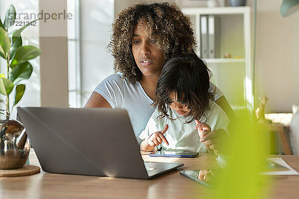 Mutter arbeitet am Laptop  während ihre Tochter mit dem Smartphone im Heimbüro spielt