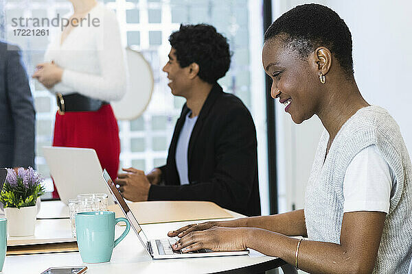 Lächelnde Geschäftsfrau arbeitet am Laptop mit Kollegen im Hintergrund im Büro