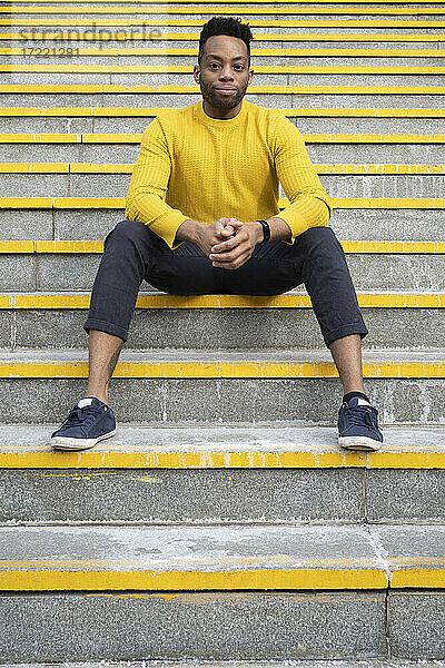 Ernster junger Mann mit gefalteten Händen auf einer gelben Treppe sitzend
