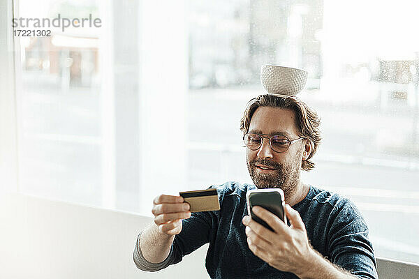 Mann balanciert Schüssel auf dem Kopf  während er in einem Café mit Kreditkarte und Smartphone bezahlt