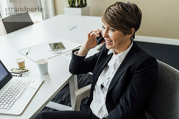 Zufriedene Unternehmerin schaut weg  während sie im Büro mit ihrem Smartphone telefoniert