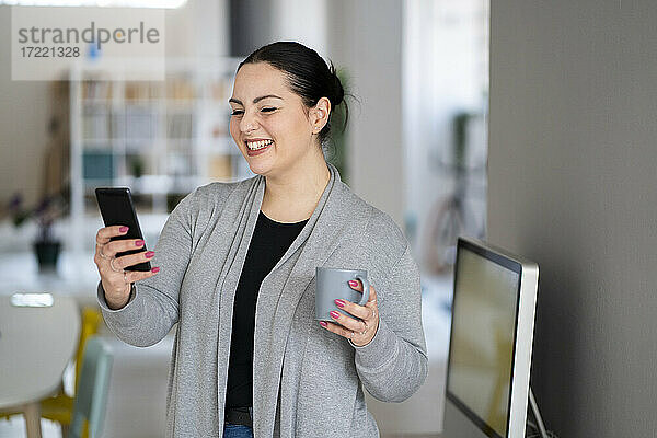 Lächelnde Geschäftsfrau mit Kaffeetasse und Telefon bei der Arbeit zu Hause