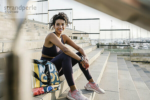 Lächelnde Sportlerin  die mit einer Tasche auf einer Treppe sitzt und träumt