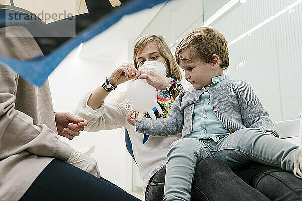 Junge spielt mit aufblasbarem Schutzhandschuh  während er mit seiner Mutter beim Zahnarzt in der Klinik sitzt