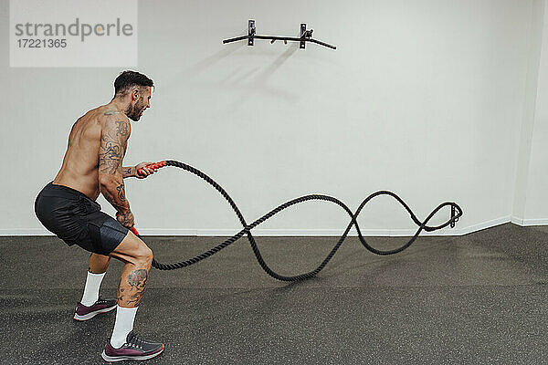Mittlerer erwachsener männlicher Sportler  der im Stehen im Fitnessstudio mit einem Seil trainiert