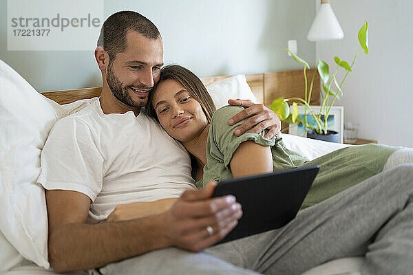 Lächelndes junges Paar  das ein digitales Tablet benutzt  während es zu Hause auf dem Bett liegt