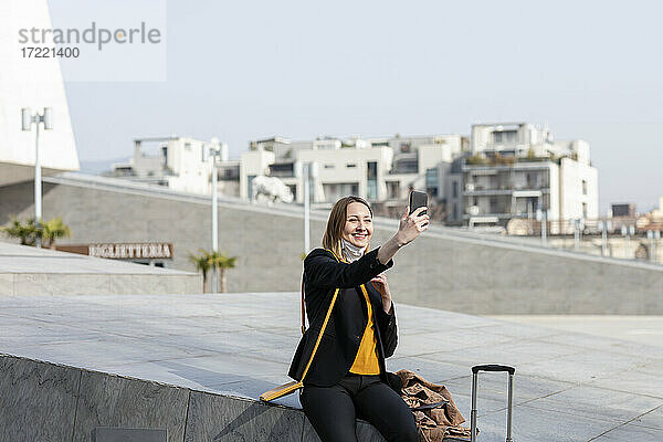 Lächelnde Geschäftsfrau  die ein Selfie mit ihrem Handy macht  während sie auf einer Stützmauer sitzt
