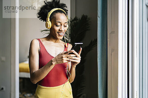 Junge Frau mit Kopfhörern  die ein Smartphone benutzt  während sie zu Hause am Fenster steht