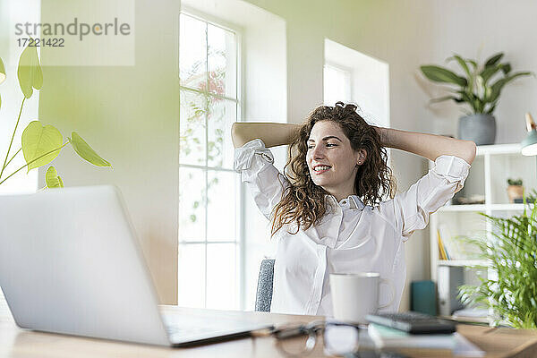 Lächelnde weibliche Fachkraft mit Händen hinter dem Kopf  die sich am Schreibtisch im Heimbüro entspannt