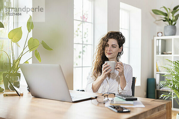 Schöne Freiberuflerin trinkt Kaffee und schaut auf den Laptop im Home Office