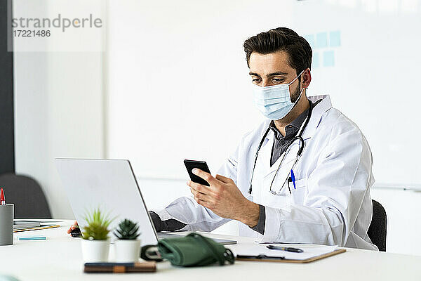 Männlicher Arzt mit Gesichtsschutzmaske und Smartphone am Schreibtisch im Krankenhaus
