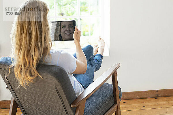 Blonde Frau hält ein digitales Tablet und entspannt sich auf einem Stuhl zu Hause