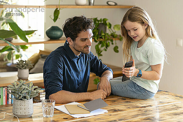 Mädchen zeigt ihrem Vater das Handy  während sie zu Hause auf dem Tisch sitzt