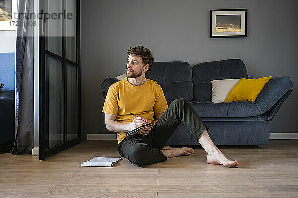 Nachdenklicher Mann hält ein Buch in der Hand und sitzt auf dem Boden im Wohnzimmer