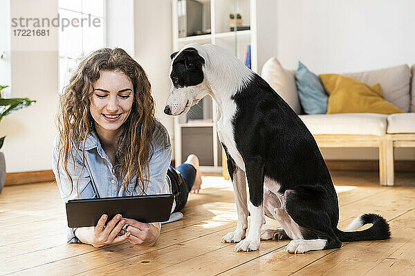 Lächelnde Frau  die auf ein digitales Tablet schaut  während sie neben einem Jack Russell Terrier im Wohnzimmer auf dem Hartholzboden liegt