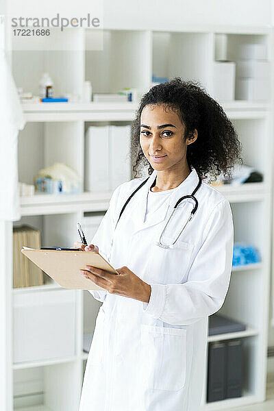 Lächelnder junger Arzt mit Klemmbrett und Stift in der Hand  während er in einer Klinik steht