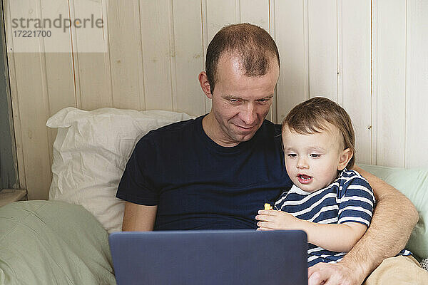Reifer Mann arbeitet am Laptop  während er mit seinem Sohn auf dem Sofa im Heimbüro sitzt
