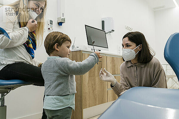 Junge spielt mit zahnmedizinischen Geräten  während er neben dem Zahnarzt und der Mutter steht  die in der Klinik eine Gesichtsmaske trägt