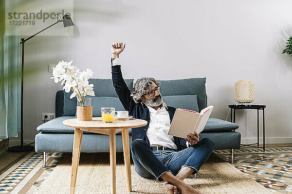 Müder älterer Mann  der seinen Arm ausstreckt  während er auf dem Sofa zu Hause ein Buch liest