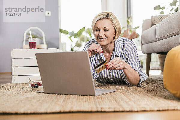 Blonde reife Frau mit Kreditkarte beim Online-Shopping mit Laptop zu Hause