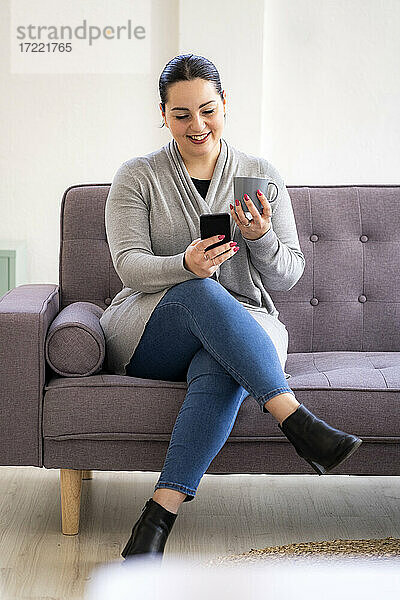 Lächelnde Frau  die ein Mobiltelefon benutzt und eine Kaffeetasse im Wohnzimmer hält